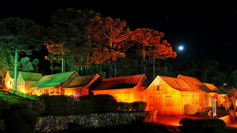 Casas típicas de madeira da serra gaúcha iluminadas à noite, sob as araucárias, em noite de lua cheia.